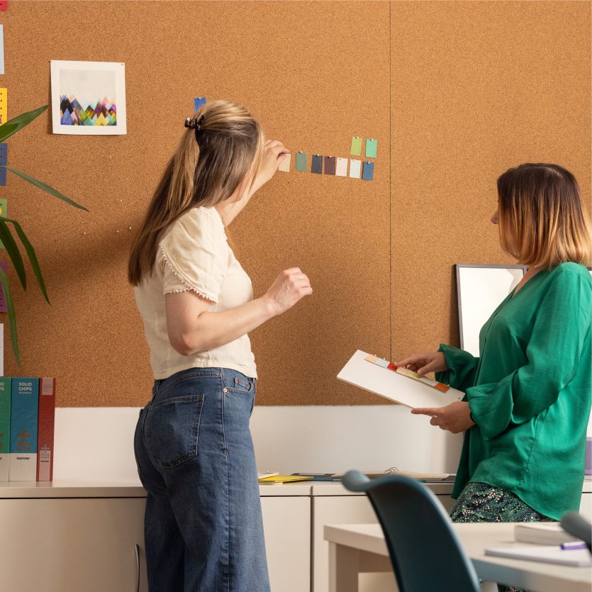 Two white females selecting colour swatches in front of a cork board in a creative office environment